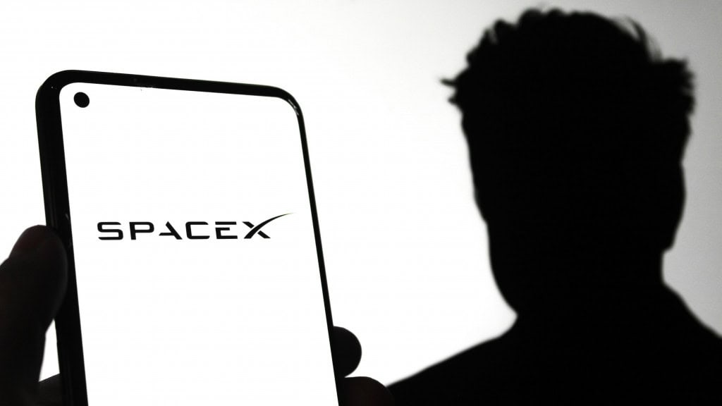 Silhueta de Elon Musk atrás de celular exibindo logomarca da SpaceX na tela