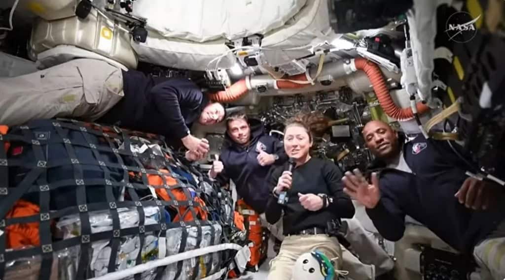 Os quatro astronautas da missão Artemis II — Jeremy Hansen, Reid Wiseman, Christina Koch e Victor Glover — flutuam no interior da cápsula Orion durante uma transmissão de vídeo. Koch segura um microfone e Glover acena para a câmera, enquanto os outros tripulantes fazem sinal de 