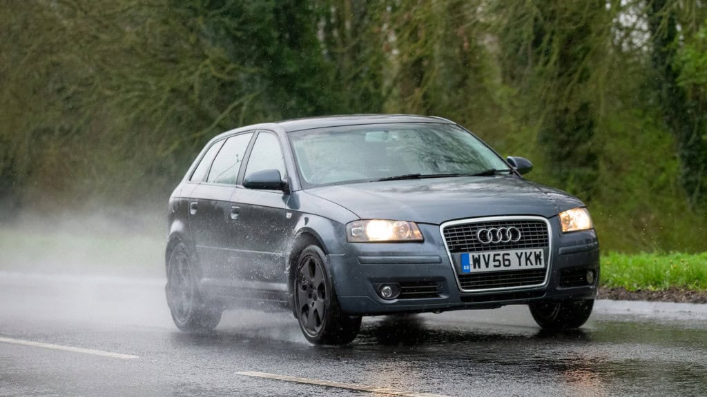 Audi A3 cinza passando por uma rua molhada