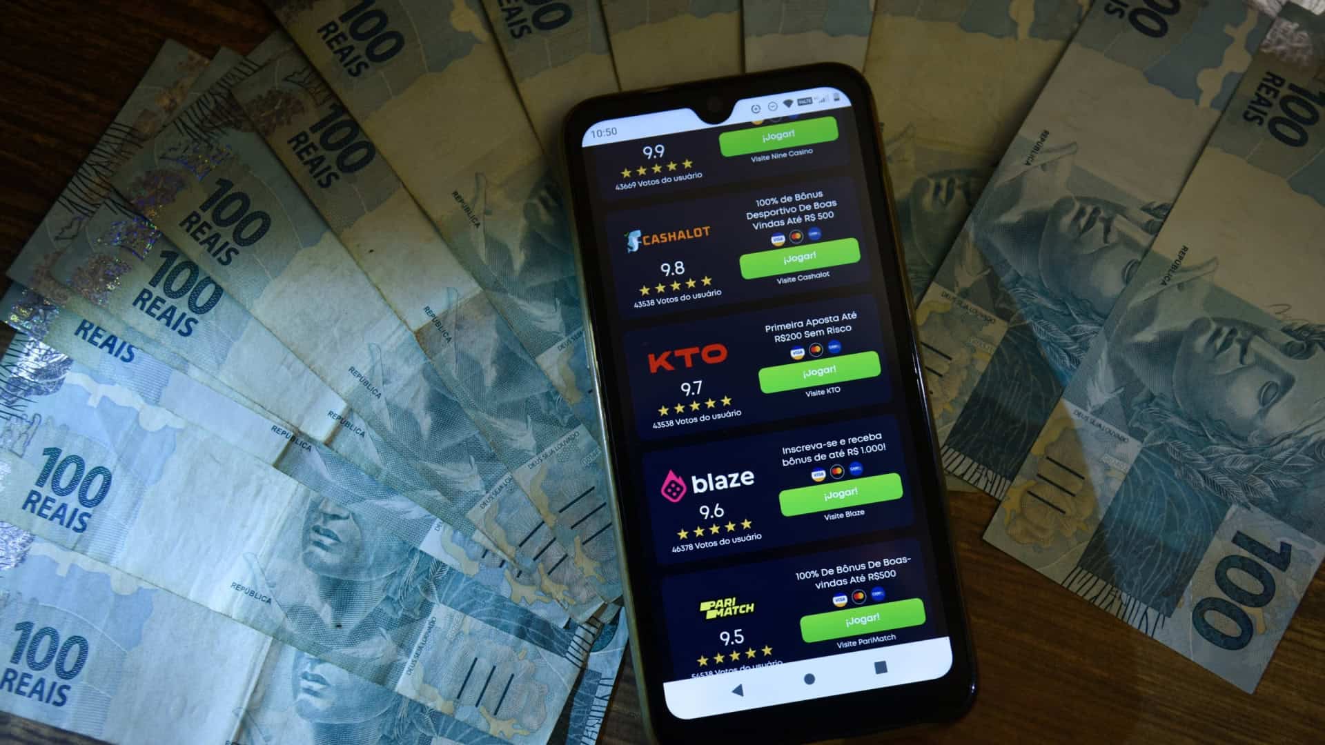 Várias notas de R$ 100 com um celular em cima, onde há o logotipo de várias bets