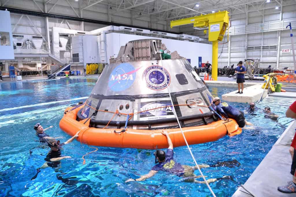 Uma versão de teste da cápsula espacial Orion, da NASA, flutua em uma piscina gigante de treinamento (Laboratório de Flutuabilidade Neutra). Diversos mergulhadores militares e equipes de resgate cercam a cápsula, que possui um anel de flutuação laranja brilhante na base. A cena ocorre dentro de um grande hangar industrial com guindastes e equipamentos de suporte