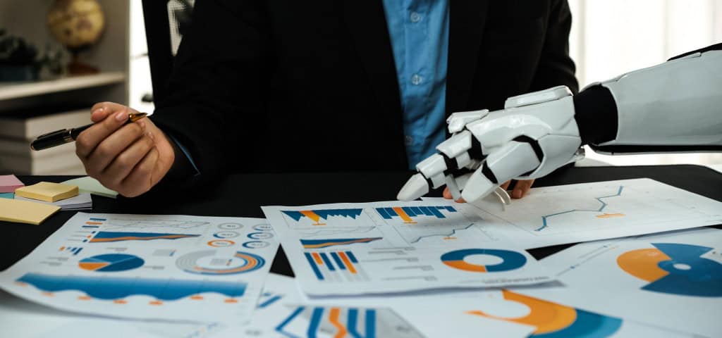 Close-up de executivo humano e braço robótico branco colaborando em mesa com relatórios de dados impressos e gráficos corporativos.