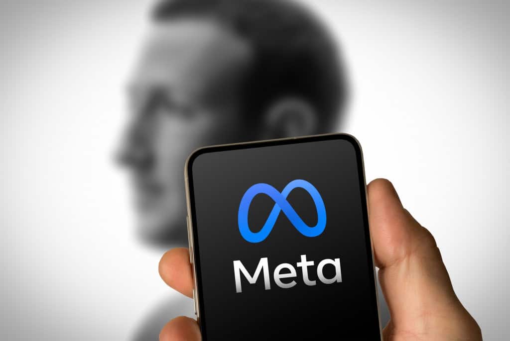 Logo da Meta exibido na tela de um smartphone, com silhueta desfocada de Mark Zuckerberg ao fundo