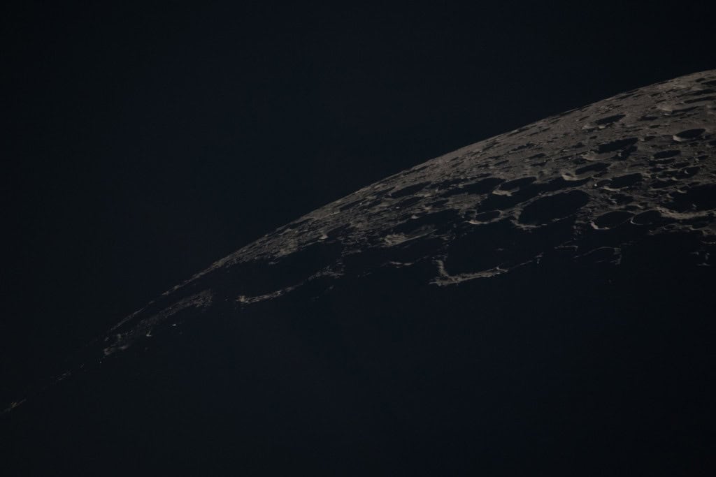 Fotografia em close-up da superfície lunar, mostrando uma paisagem repleta de crateras de diversos tamanhos em tons de cinza e sombras profundas. A curvatura da Lua corta a imagem diagonalmente contra o fundo preto.