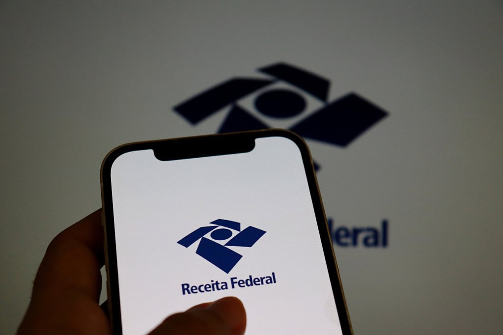 Logo da Receita Federal ao fundo e à frente, na tela de um smartphone