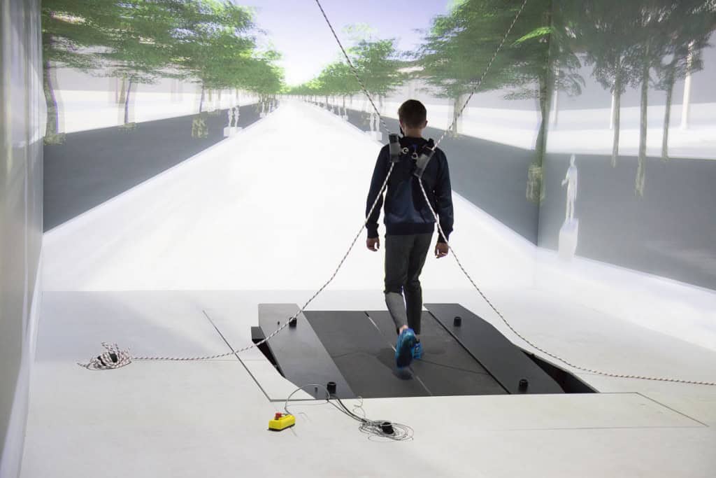 Imagem mostra um rapaz em um dispositivo similar a uma esteira ergométrica inclinada, dentro de uma sala fechada, andando