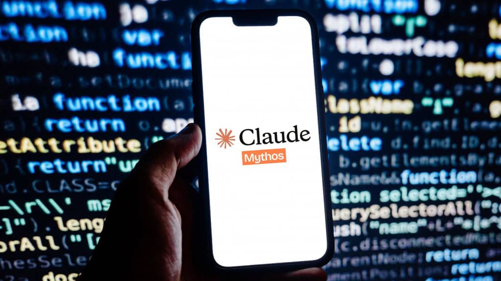 Pessoa segurando celular com logomarca do Claude Mythos na tela; ao fundo, monitor exibe linhas de código de programação