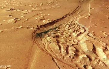 Imagem aérea de uma área de Marte com um cânion sinuoso cercado por terreno irregular e formações rochosas elevadas. A paisagem avermelhada apresenta marcas de erosão e regiões com relevo acidentado.