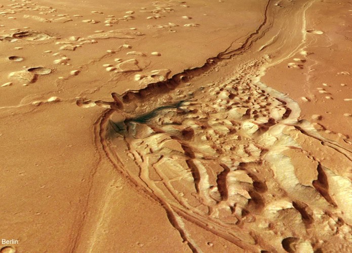 Imagem aérea de uma área de Marte com um cânion sinuoso cercado por terreno irregular e formações rochosas elevadas. A paisagem avermelhada apresenta marcas de erosão e regiões com relevo acidentado.