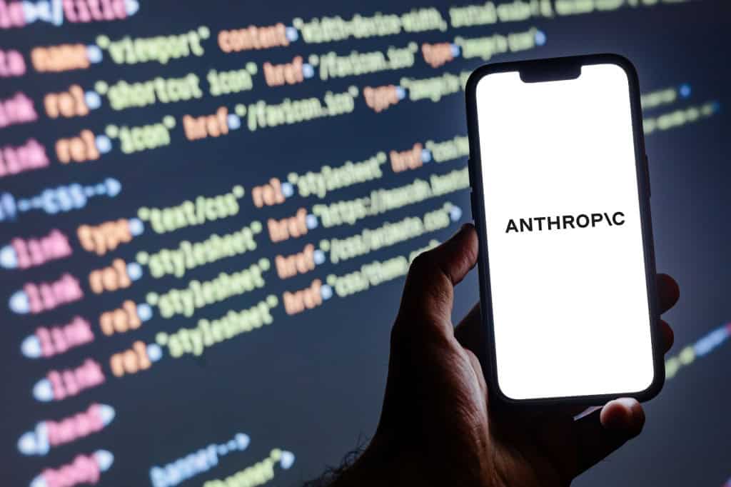 Ao fundo, linhas de código; à frente, em um celular segurado por uma mão, o logo da Anthropic