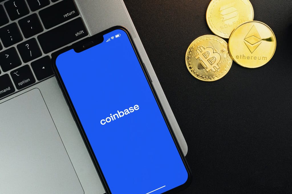 Três moedas ao lado de um notebook, o qual possui, em cima, um smartphone; nele, há o logo da Coinbase