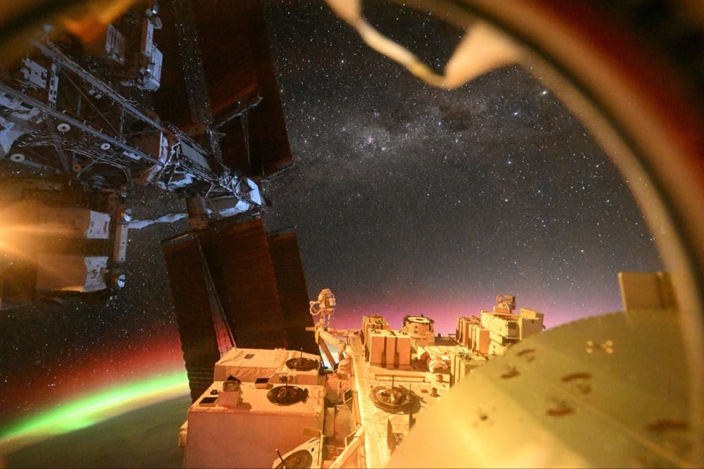 Vista da Estação Espacial Internacional mostra auroras verdes e vermelhas sobre a Terra, com estrelas e partes da estrutura da ISS em primeiro plano