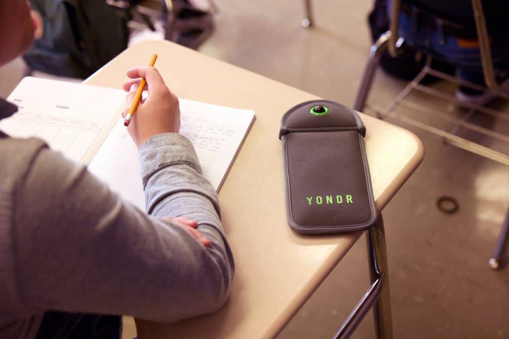 Imagem de divulgação da fabricante Yondr mostrando uma bolsa armazenamento um celular em cima de mesa escolar.