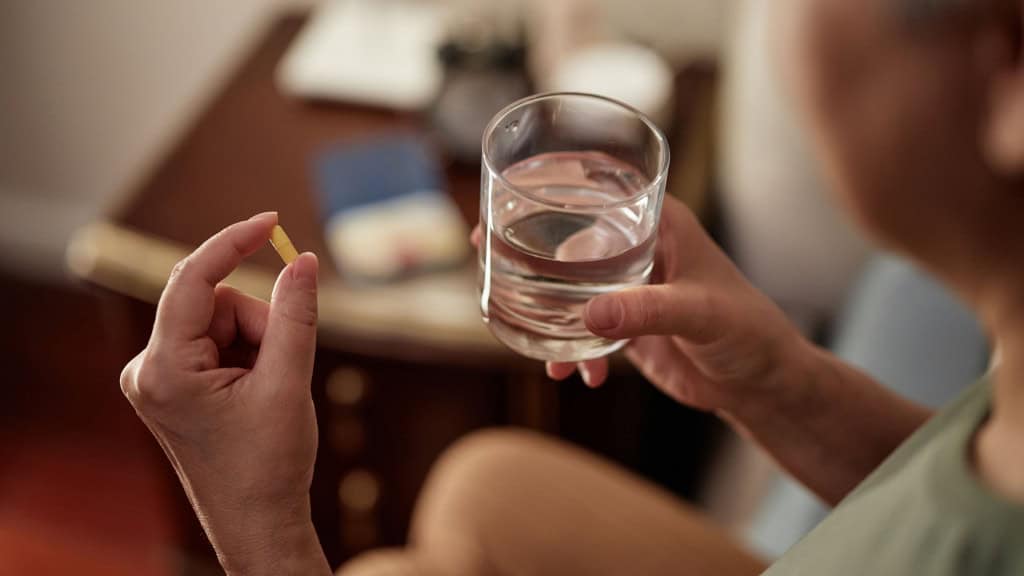 Mulher idosa segurando cápsula de remédio com uma mão e copo de água com outra
