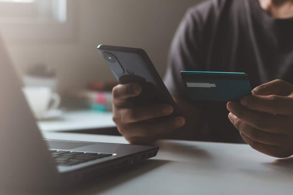 Pessoa segura um smartphone e um cartão bancário ao realizar uma compra online em frente a um notebook