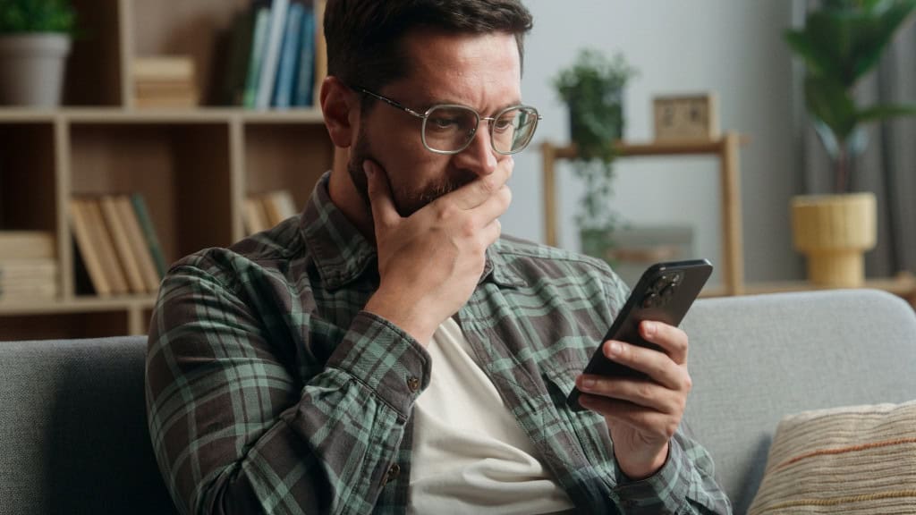 Homem olha preocupado para a tela de um smartphone enquanto está sentado em um sofá