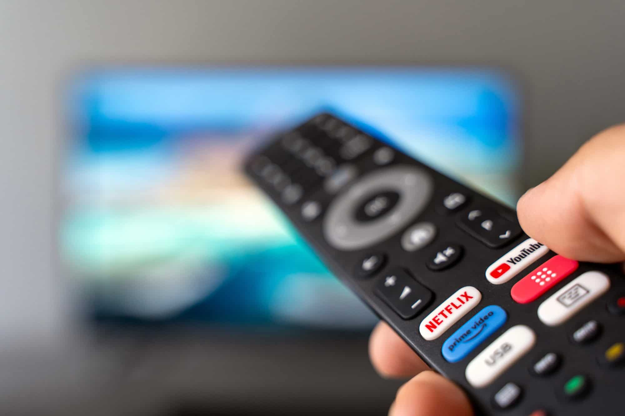 Controle remoto apontado para uma televisão com interface de streaming desfocada ao fundo.