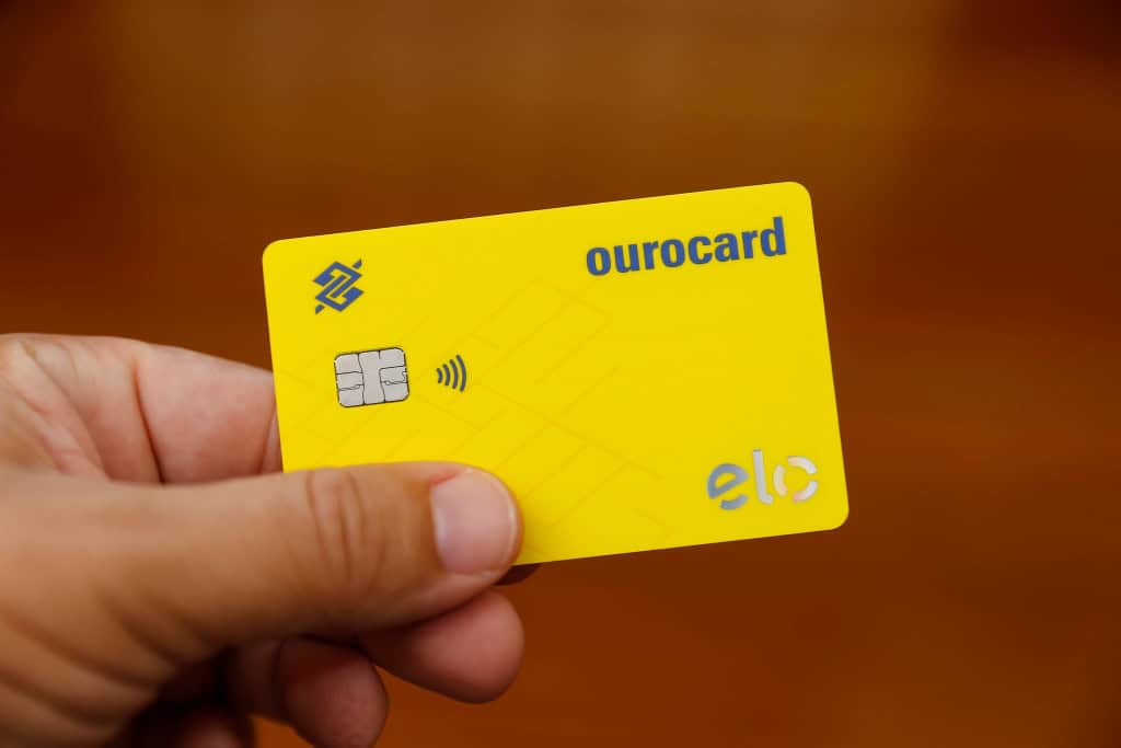 Cartão de crédito da bandeira Elo sendo segurado por uma pessoa, em destaque sobre fundo desfocado em tons alaranjados