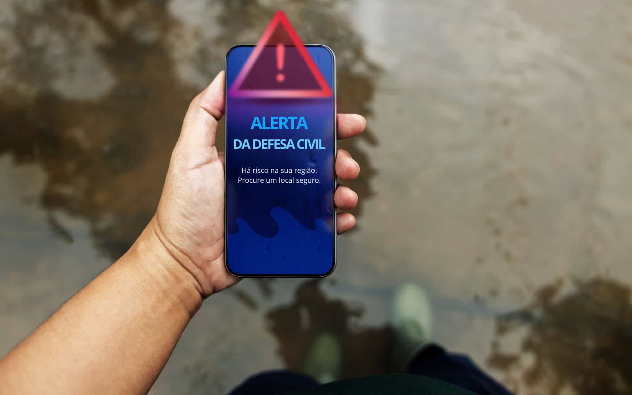 Celular exibe alerta da Defesa Civil enquanto pessoa caminha por uma área alagada.