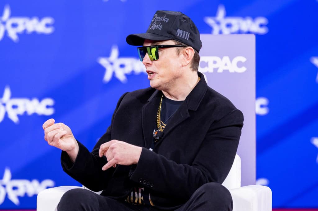 Elon Musk de óculos e boné, sentado e gesticulando