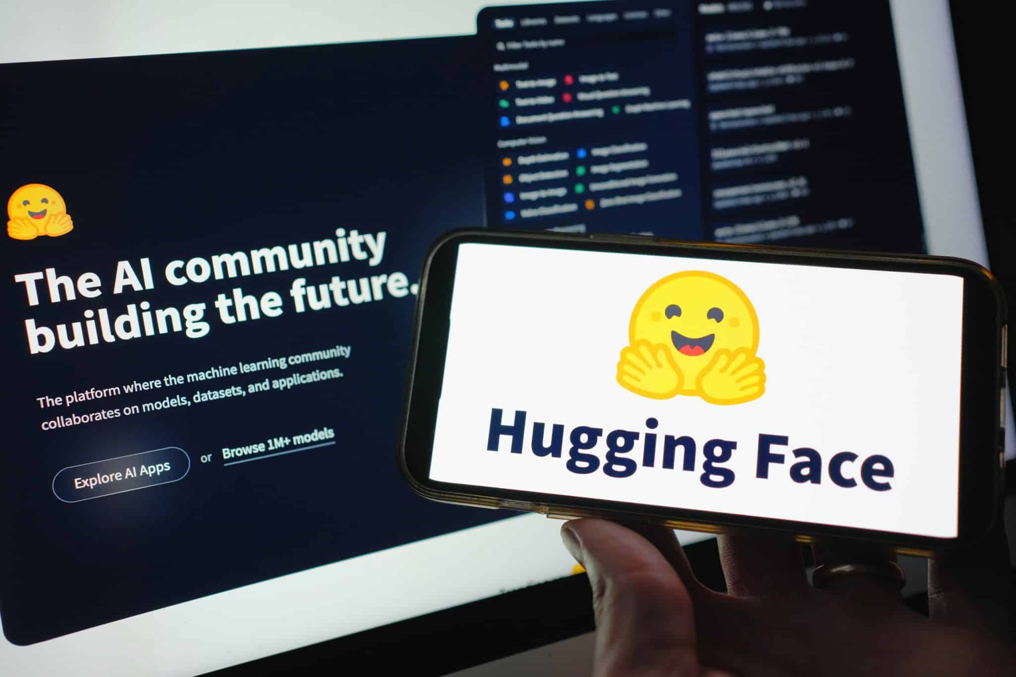 Página inicial do Hugging Face ao fundo; à frente, logo da empresa em um smartphone que está na mão de uma pessoa