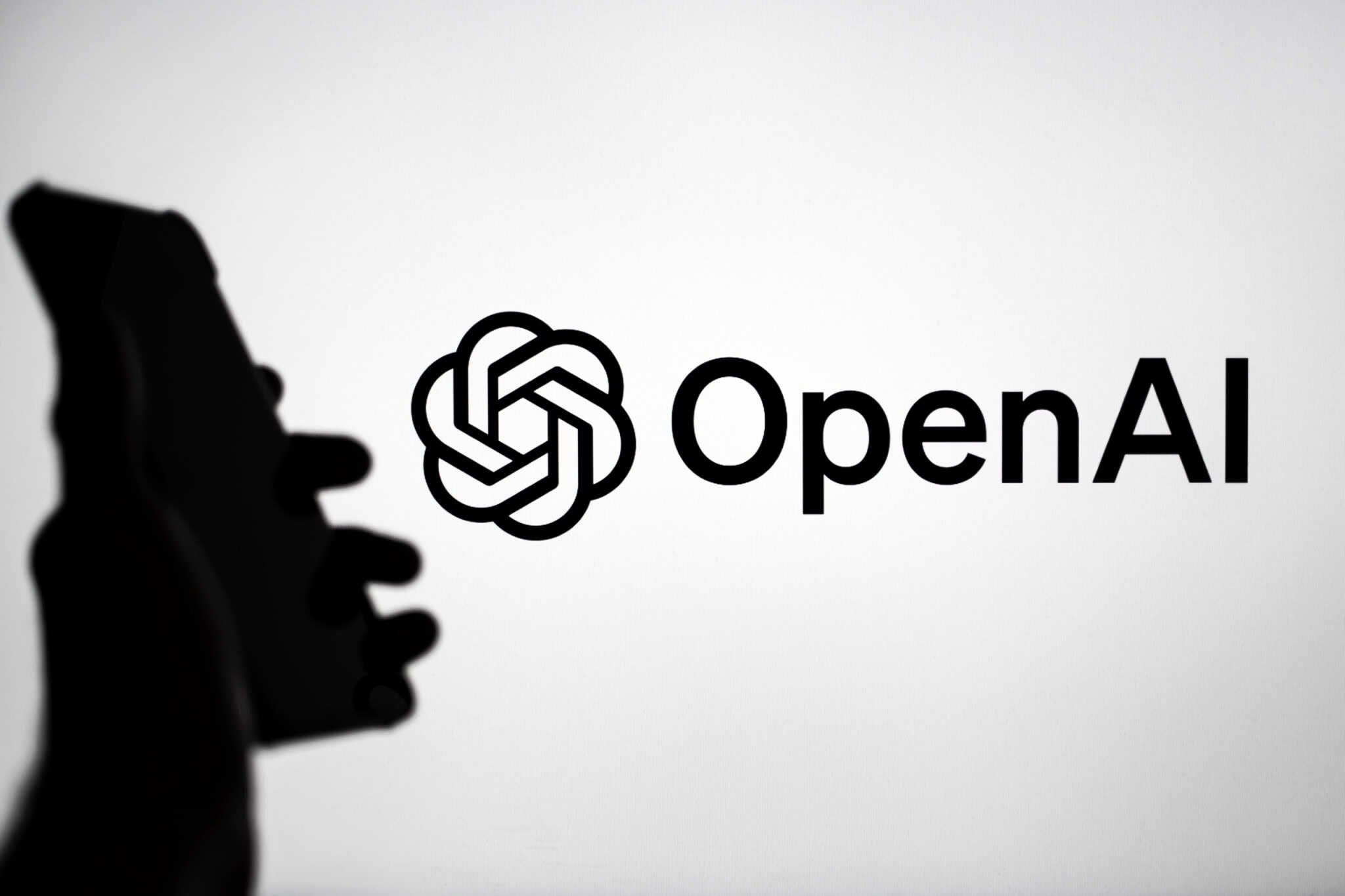 Ao fundo, logo da OpenAI; à frente e a esquerda, sombra de uma mão segurando um smartphone