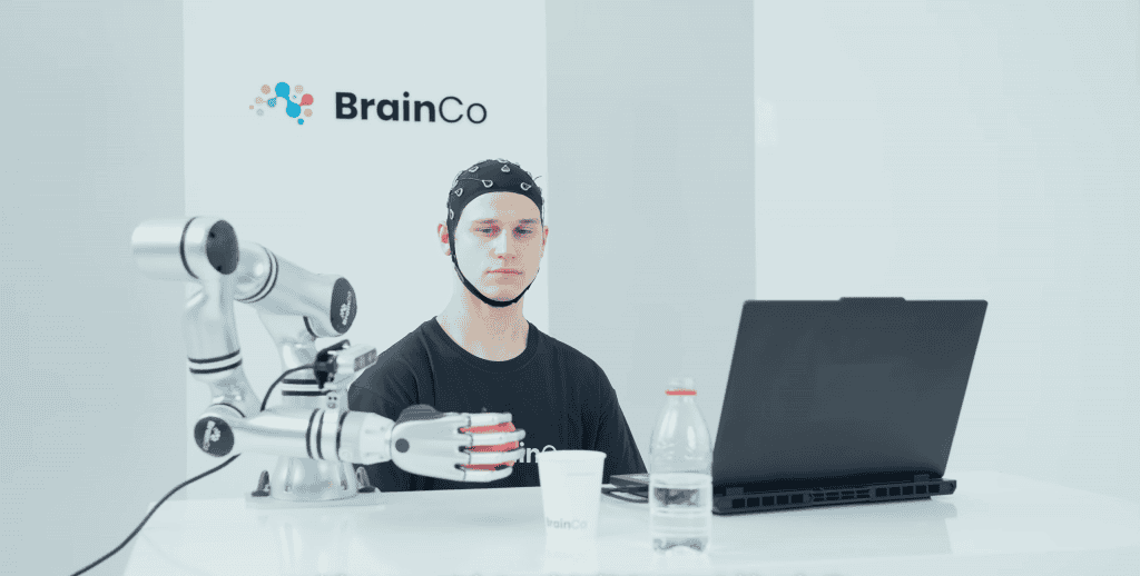 Pessoa controlando mão de robô em frente a um notebook; na mão de robô, há um copo