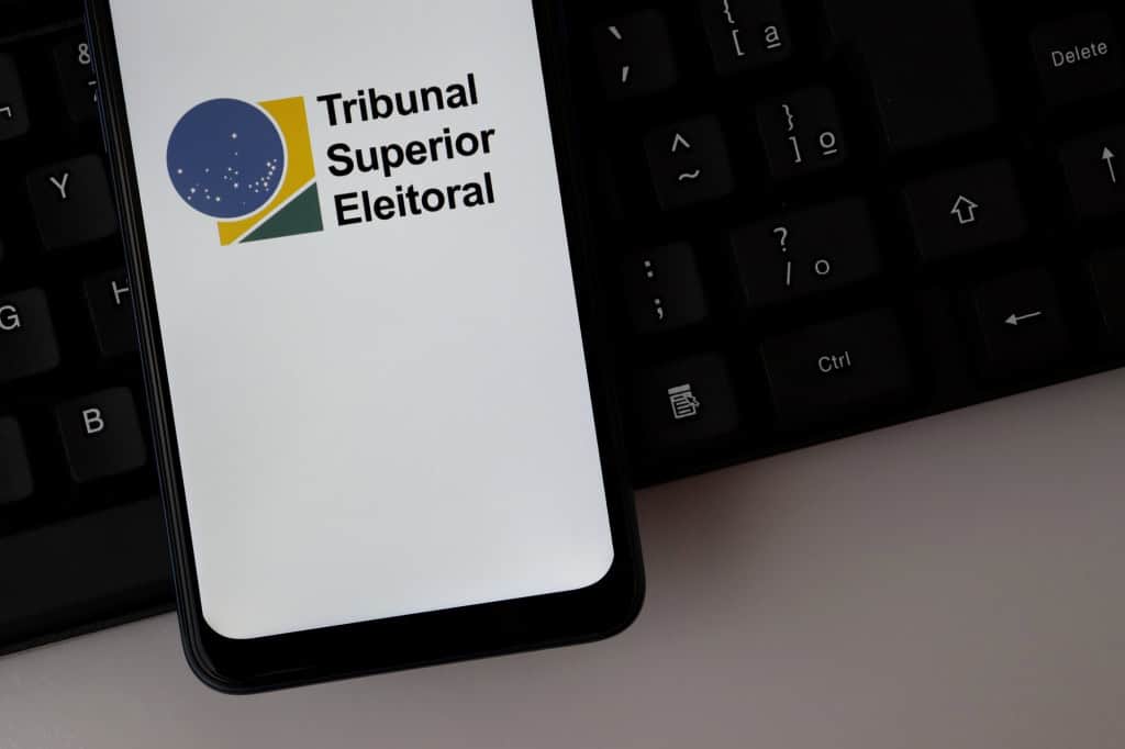 Logotipo do Tribunal Superior Eleitoral (TSE) exibido na tela de um smartphone sobre um teclado de computador.