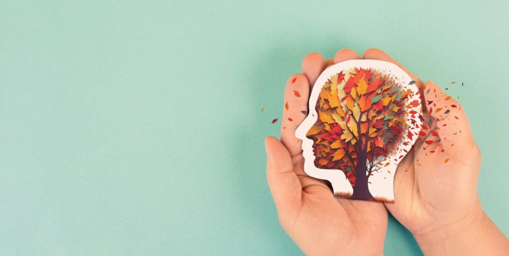 uma mão segurando um desenho de uma cabeça com o cérebro representado por uma árvore sob o vento para demonstrar as condições de um paciente com Alzheimer.