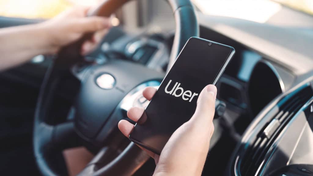 Pessoa segurando volante de carro com uma mão e celular com a outra; tela do celular exibe logomarca da Uber