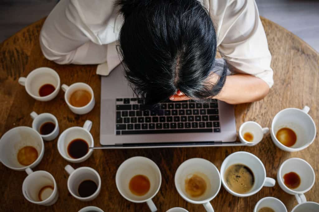 Diversas xícaras de café sobre uma mesa ao lado de uma pessoa apoiada em um notebook.