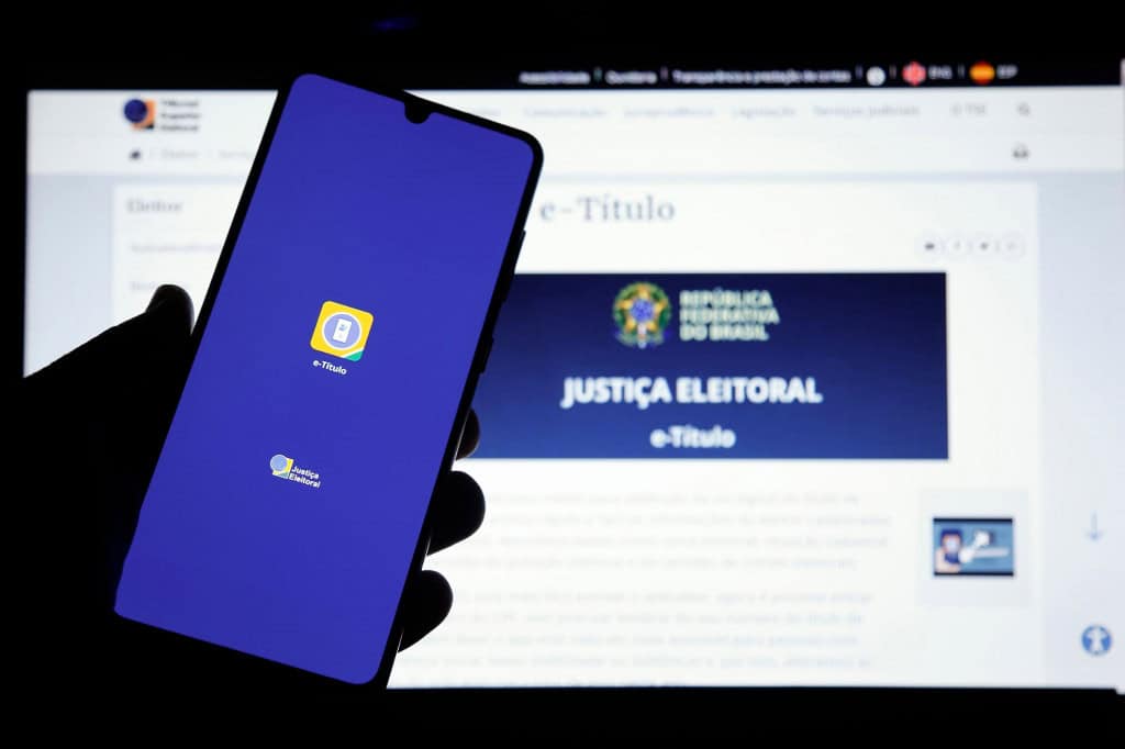 Homem segurando um celular com a tela do aplicativo e-Título sobre um monitor com o site do e-Título.