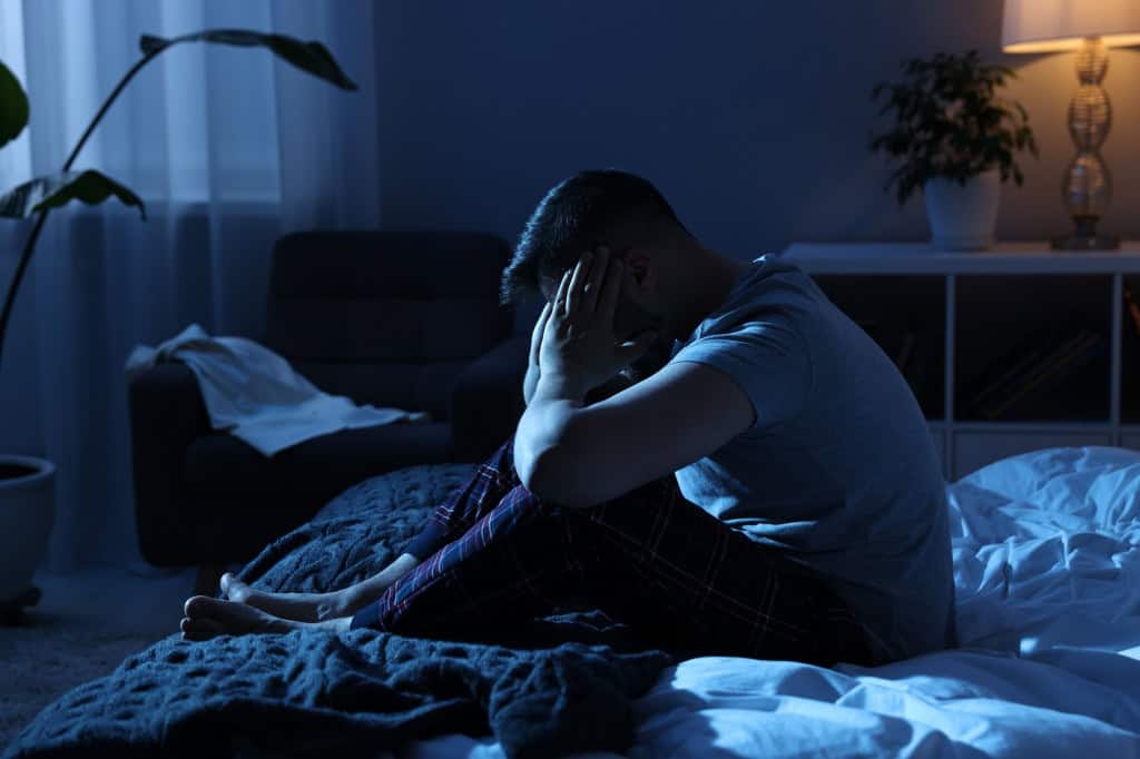 Homem sentado na cama durante a madrugada, com as mãos no rosto, após acordar no meio da noite.
