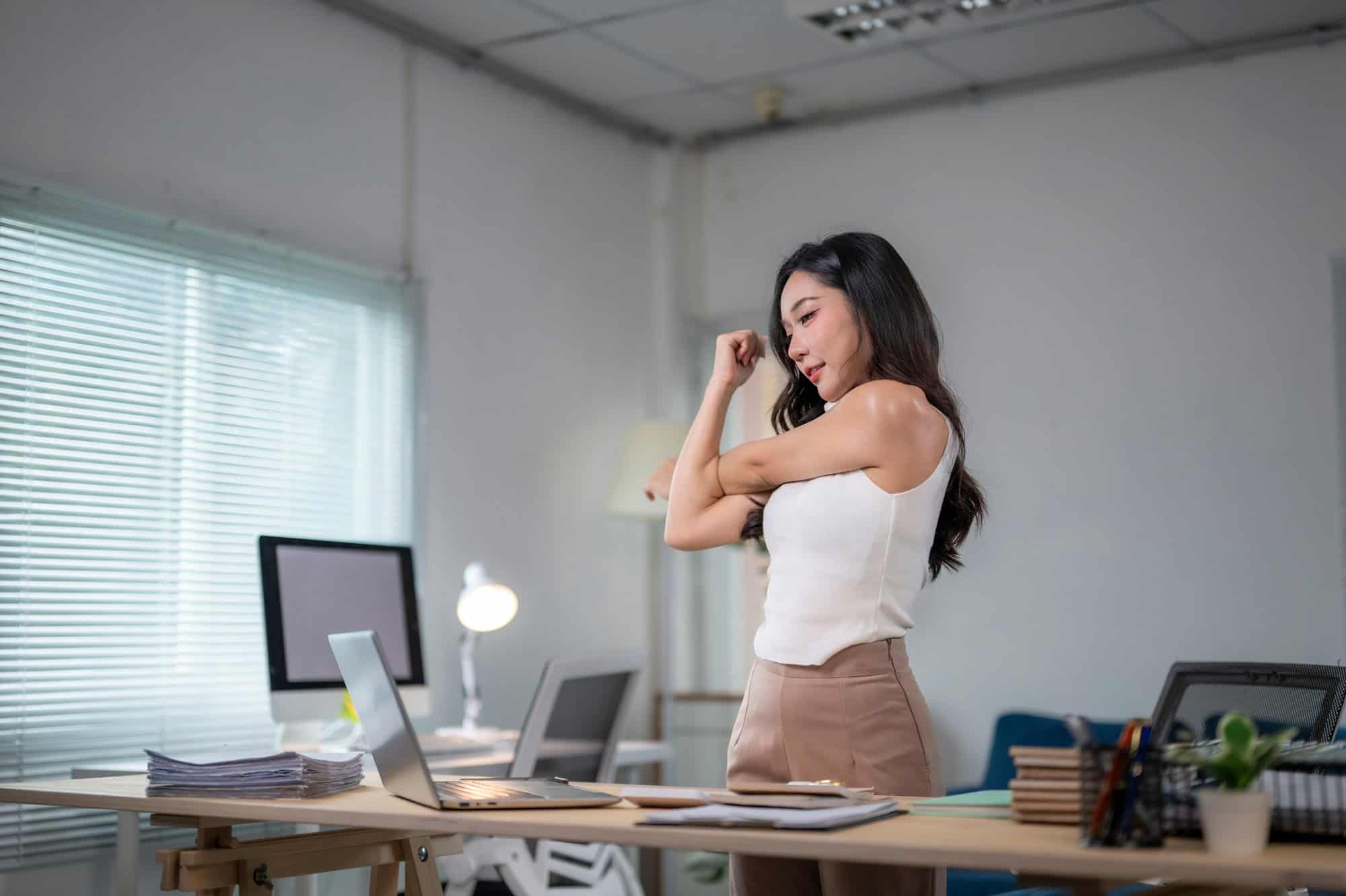 Mulher em pé alongando os braços em um escritório ao lado de uma mesa com computador.