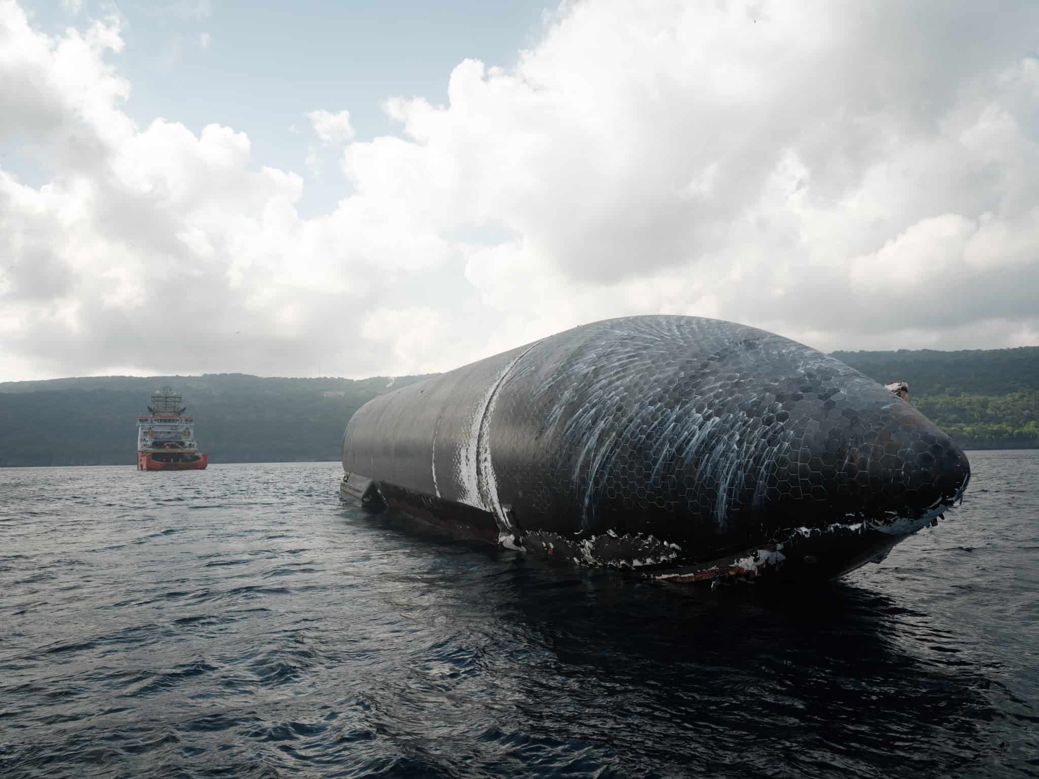 Starship parcialmente submersa no oceano, com o escudo térmico danificado, enquanto um navio de apoio se aproxima ao fundo