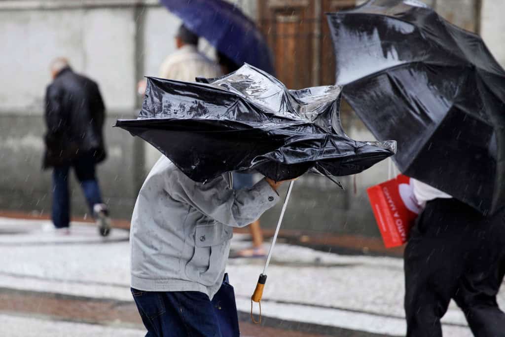 Pessoa segura um guarda-chuva virado pela força do vento durante um temporal, enquanto outras caminham sob chuva ao fundo.