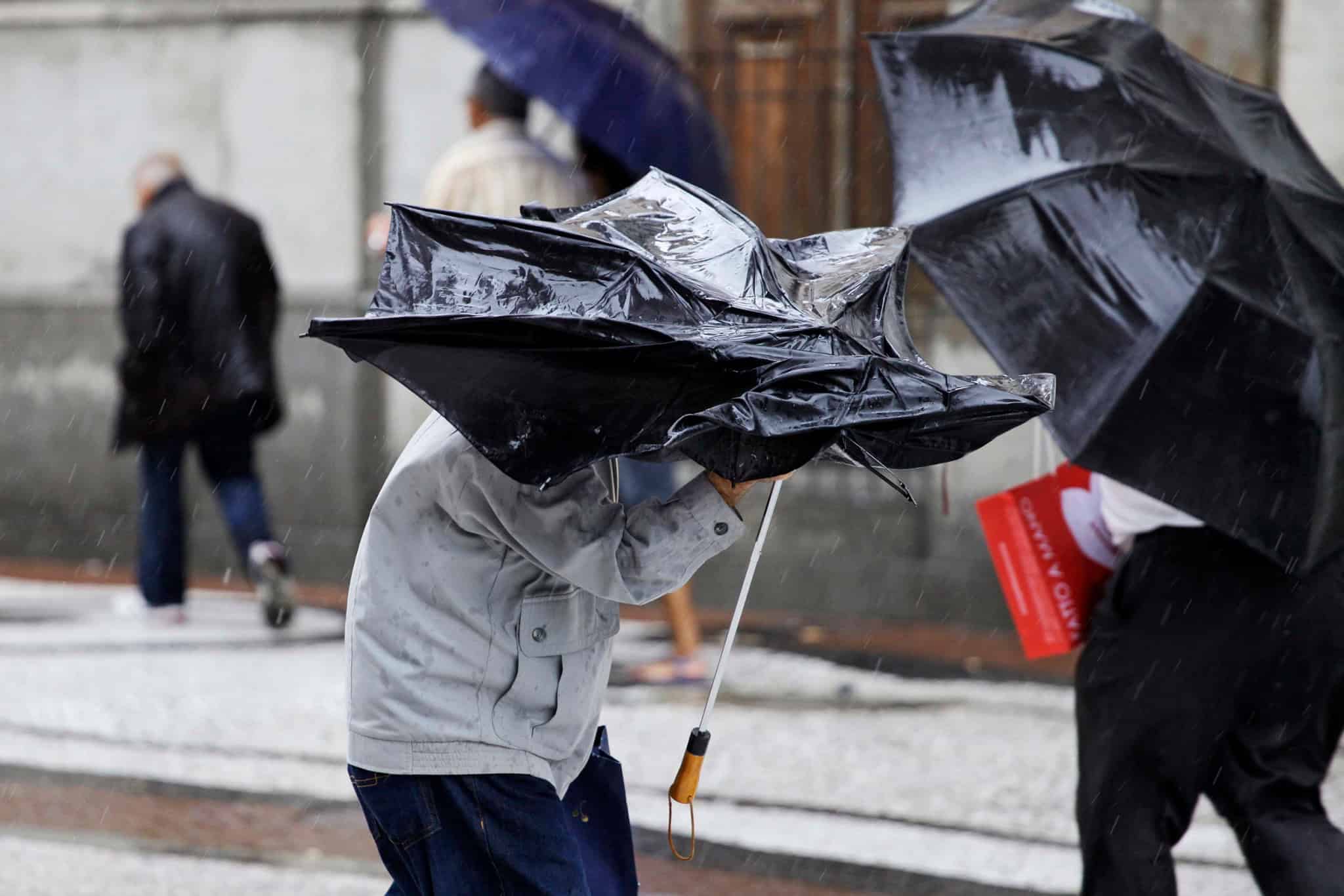 Pessoa segura um guarda-chuva virado pela força do vento durante um temporal, enquanto outras caminham sob chuva ao fundo.