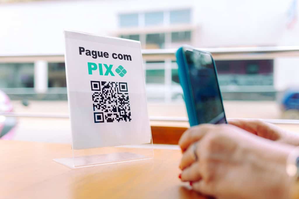 Imagem editorial ilustrativa: mãos segurando um smartphone e escaneando um código QR em um suporte de acrílico para pagamento via PIX, sobre uma mesa de bar ao ar livre, com uma rua da cidade ao fundo.