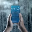 Mãos segurando um smartphone mostrando um aplicativo de previsão do tempo da chuva contra uma janela coberta de gotas de chuva em um dia frio e chuvoso
