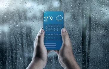 Mãos segurando um smartphone mostrando um aplicativo de previsão do tempo da chuva contra uma janela coberta de gotas de chuva em um dia frio e chuvoso