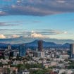Vista panorâmica da Cidade do México, com edifícios e áreas urbanizadas em primeiro plano e montanhas com picos nevados ao fundo, sob um céu parcialmente nublado.