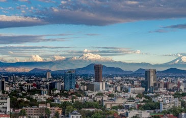 Vista panorâmica da Cidade do México, com edifícios e áreas urbanizadas em primeiro plano e montanhas com picos nevados ao fundo, sob um céu parcialmente nublado.