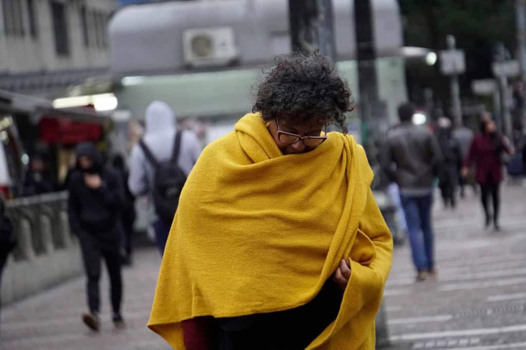 Pessoa enrolada em manta caminha em rua durante chegada de frente fria em São Paulo