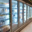 Corredor de supermercado com geladeiras e freezers expositores alinhados, com portas de vidro e produtos refrigerados nas prateleiras.