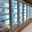 Corredor de supermercado com geladeiras e freezers expositores alinhados, com portas de vidro e produtos refrigerados nas prateleiras.