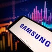Logo da Samsung em um smartphone; ao fundo, gráficos financeiros